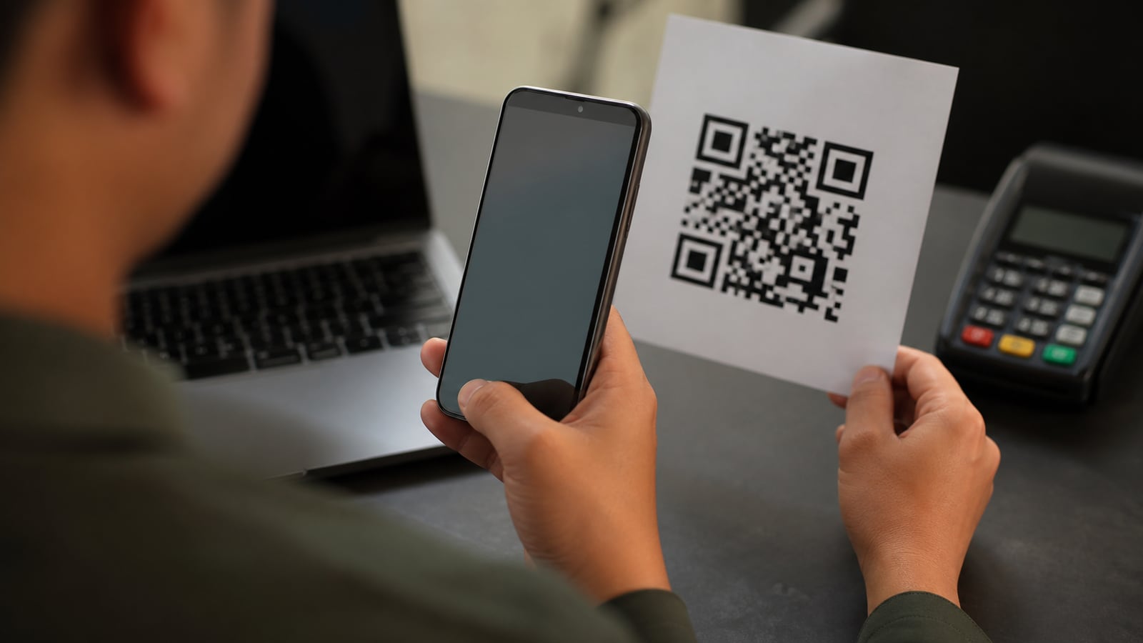 Persoon controleert een QR-code met smartphone naast betaalterminal en laptop